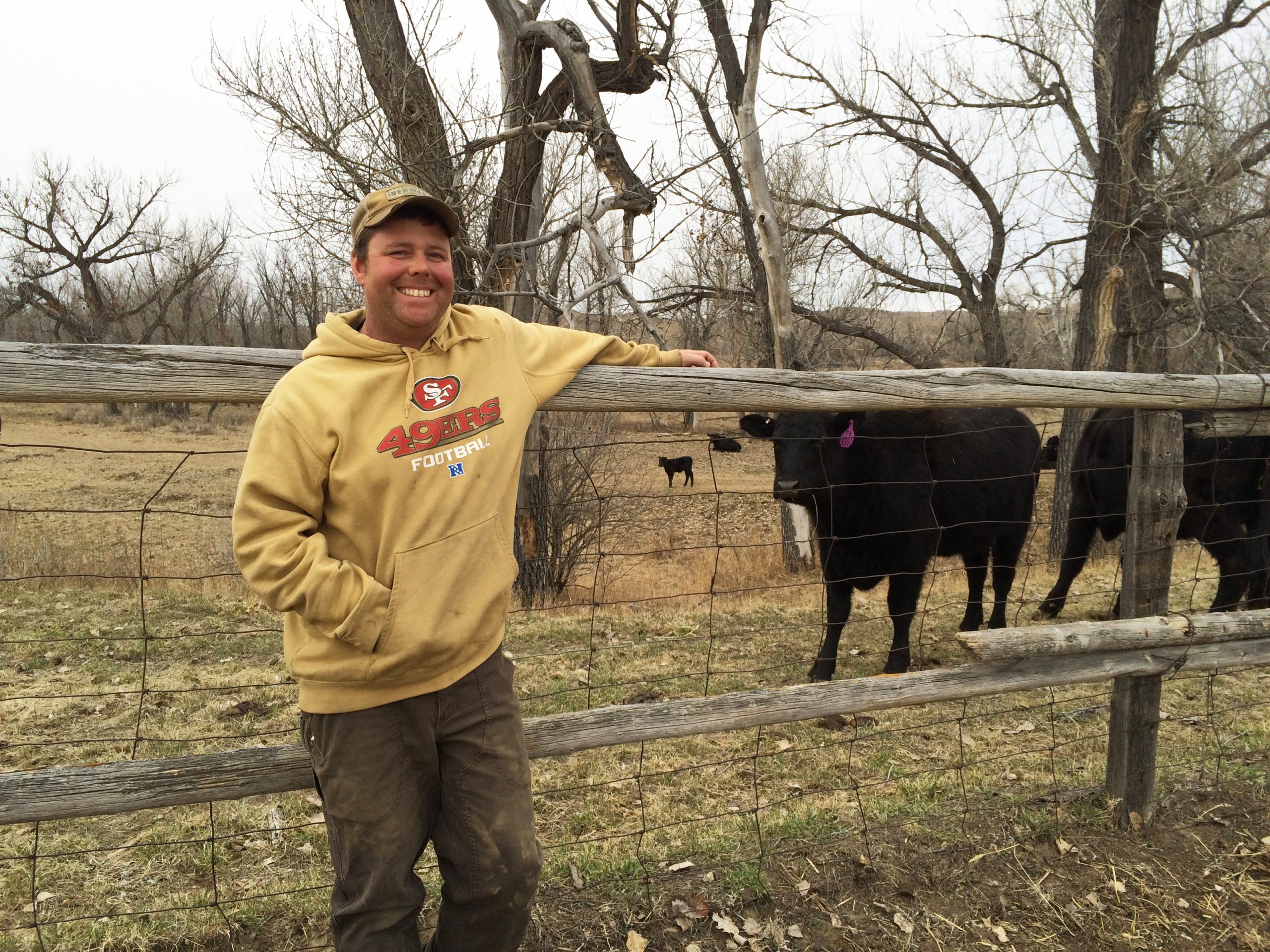 Man in a tan hoodie, brown baseball cap, and dark brown pants, leaning on a fence in front of a black cow outside